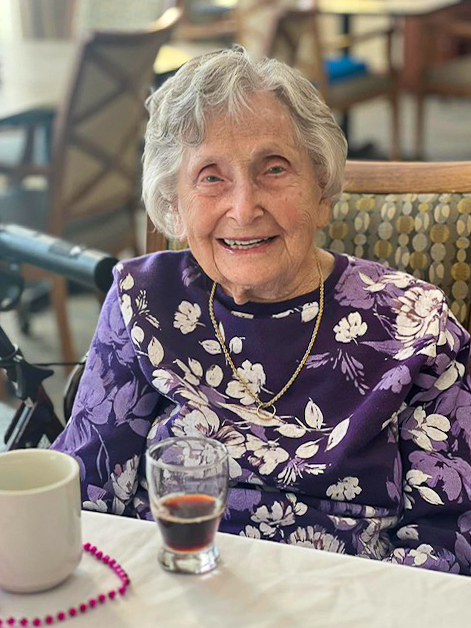 A recent photo of Julia at the Cedar Trails Community, sitting down to dine in her purple blouse with detailed flowers, radiates grace and warmth.