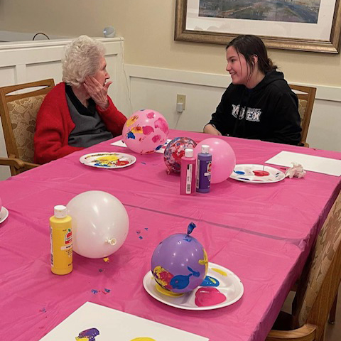 Two women having fun at a table, painting with balloons. A young lady bonds with a memory care resident in the neighborhood.