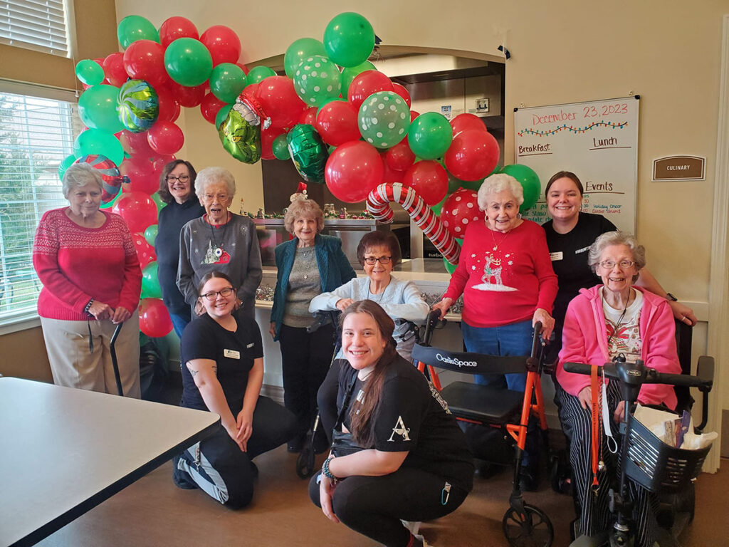 In the spirit of celebration, a cheerful group of employees and residents at Cedar Trails Senior Living pose happily in front of a Holiday arch adorned with red and green balloons.