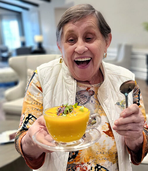 Smiling senior resident joyfully holds a bowl of vibrant orange soup garnished with greens, wearing a colorful butterfly-patterned shirt and white vest, ready to enjoy the meal.