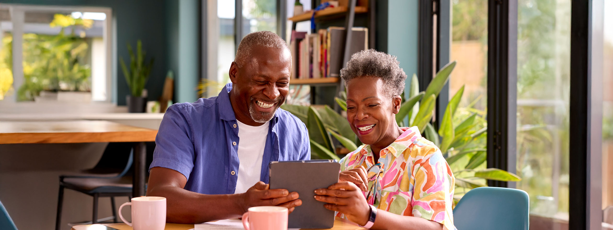 Joyful senior couple sitting at a kitchen table, laughing while using a tablet together, enjoying technology and quality time in a bright, plant-filled home.