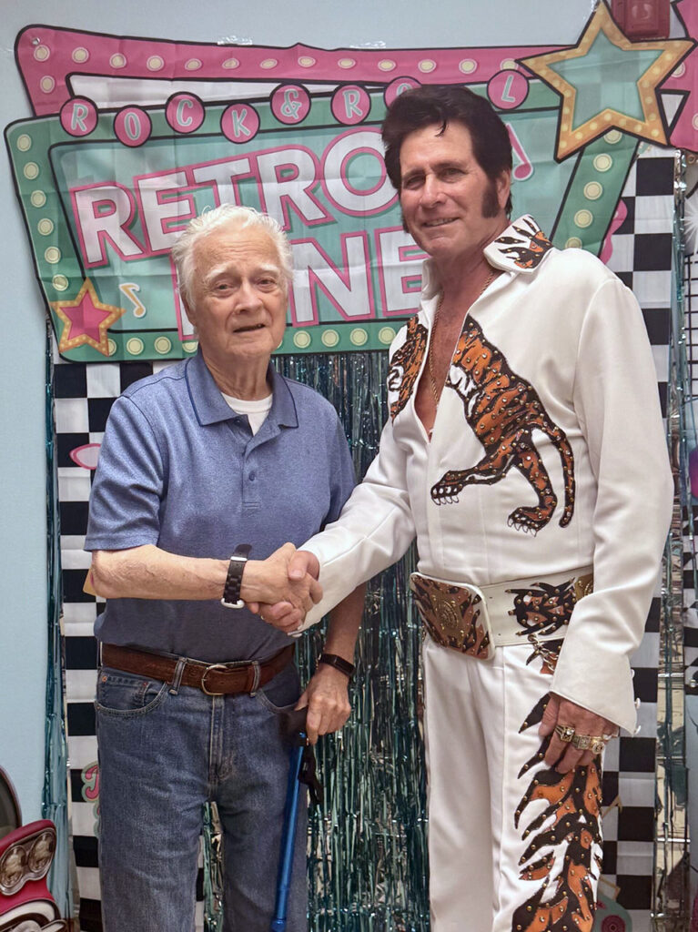 Senior resident shakes hands with Elvis impersonator at a retro diner-themed event, celebrating music, nostalgia, and joyful moments together.
