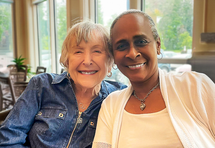 Two smiling women sit closely together by a window, radiating warmth and friendship during a cheerful moment in their senior living community.