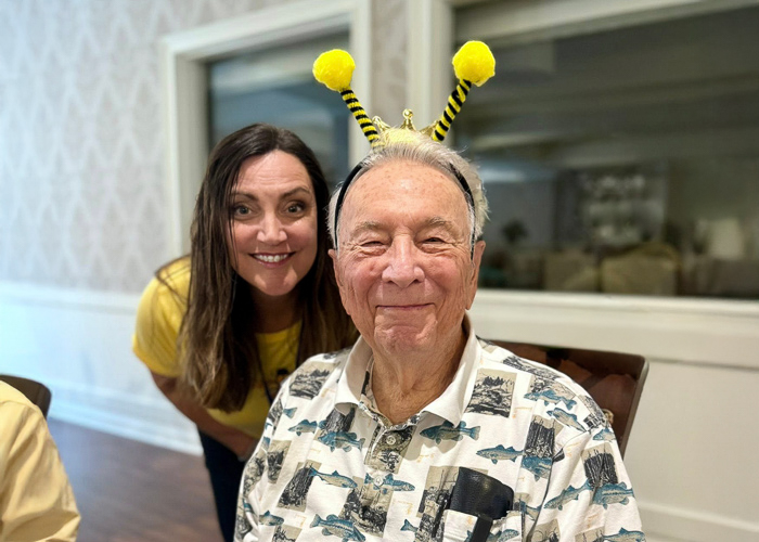 A senior resident wearing playful bee antenna headbands smiles with joy while a team member leans in beside him, celebrating a fun themed event in the community.
