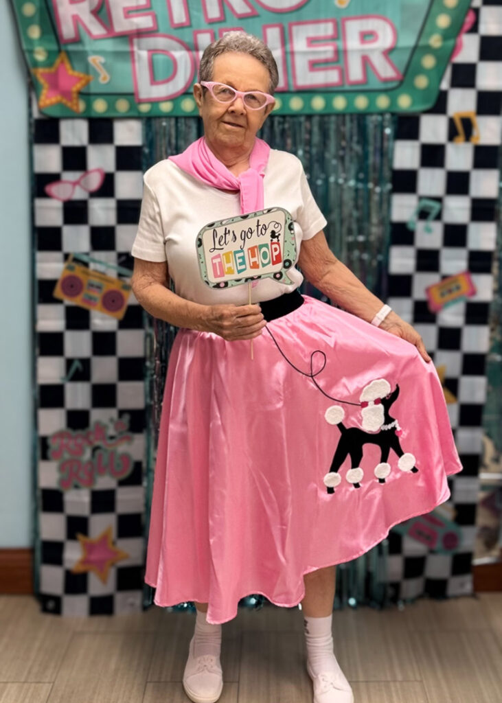 A senior woman dressed in a retro outfit poses in front of a "Retro Diner" backdrop, holding out a pink skirt with poodle on it.