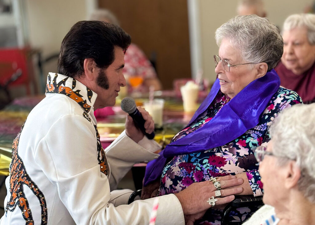 An Elvis impersonator sings to an attentive senior woman, with other residents enjoying the performance in the background.
