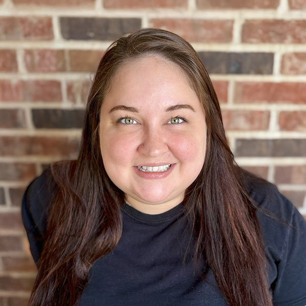 Ashley Mazotas, Memory Care Director at Cedar Trails Senior Living, smiling in a professional headshot. She has long brown hair, wears a black top, and stands in front of a red and beige brick wall background.