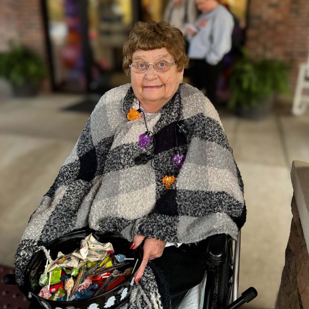 A senior woman wrapped warmly in plaid smiles, holding a bucket of Halloween candy for trick-or-treaters.