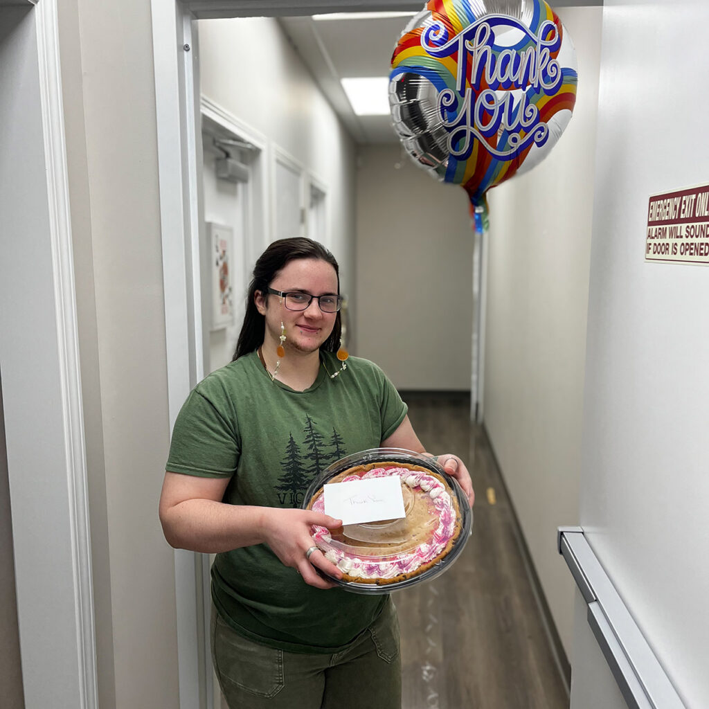A local business owner smiles, holding a balloon, large decorated cookie, and card, given in thanks.