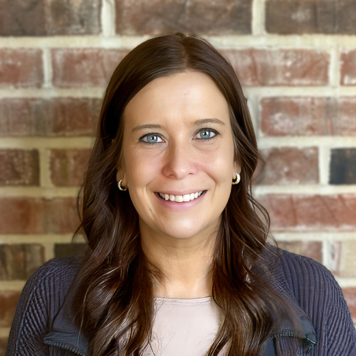 Gina Mesnarich, Memory Care Director at Cedar Trails Senior Living, smiling in a professional headshot with long dark wavy hair, blue eyes, gold hoop earrings, and a red brick wall background.