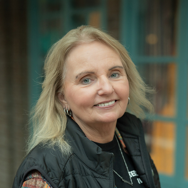Lori Peterson, Resident Services Director at Cedar Trails Senior Living, smiling in a professional outdoor headshot with light blonde hair, silver hoop earrings, a black vest, and softly blurred teal window details in the background.