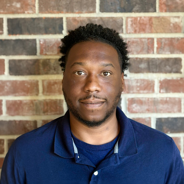 Mark Murphy, Plant Operations Director at Cedar Trails Senior Living, looking at the camera in a professional headshot with short curly hair, a trimmed beard, a navy polo shirt, and a red brick wall background.
