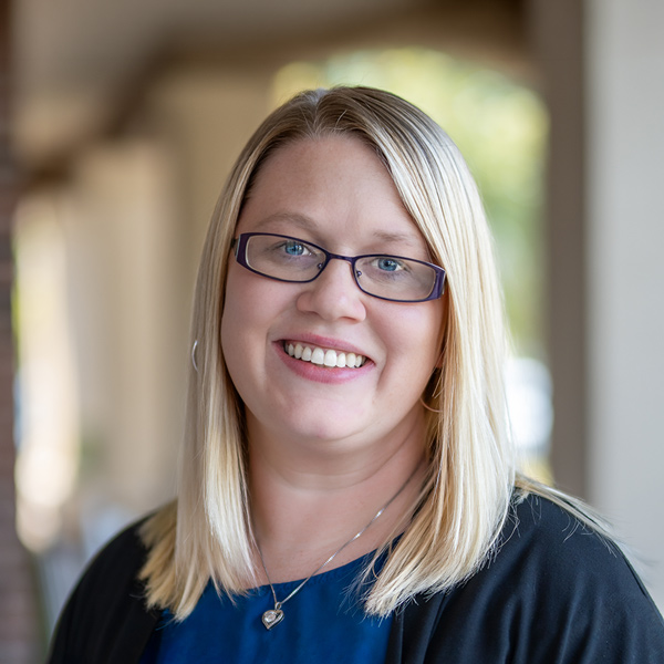 Brianna “Bri” Wilson, Business Operations Director at Cedar Trails Senior Living, smiling in a professional headshot with straight blonde hair, blue rectangular glasses, a blue top, and a softly blurred outdoor background.