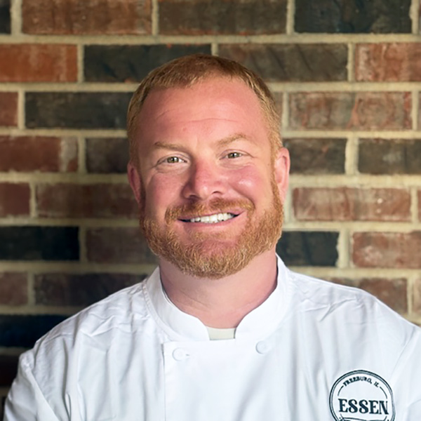 Justin Hewitt, Culinary Director at Cedar Trails Senior Living, smiling in a professional headshot with short red hair, a full ginger beard, a white chef coat, and a red brick wall background.