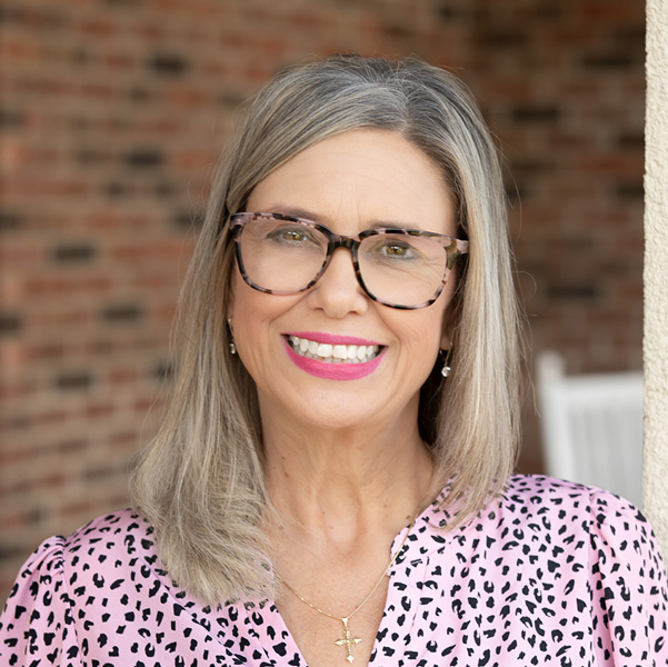 Kim Chase, Executive Director at Cedar Trails Senior Living, smiling in a professional headshot with shoulder-length blonde-gray hair, oversized patterned glasses, bright pink lipstick, a pink patterned blouse, and a softly blurred brick background.