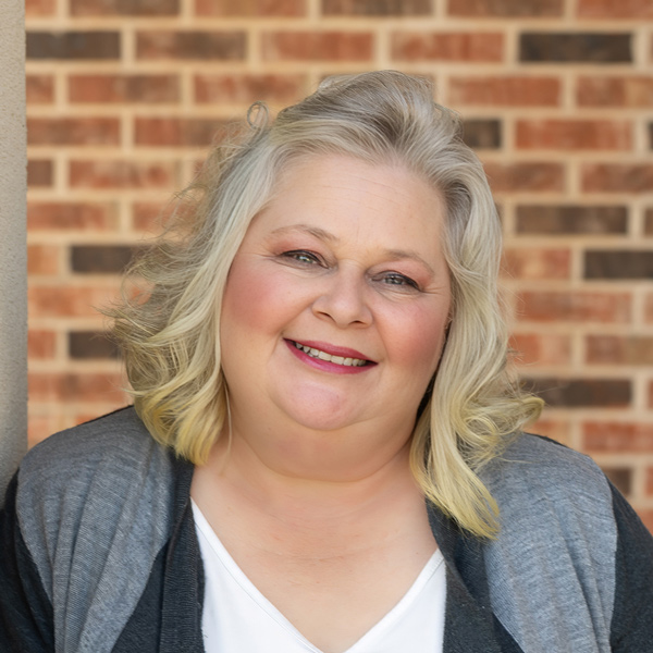 Misty Johnson, Senior Living Director at Cedar Trails Senior Living, smiling in a professional headshot with shoulder-length wavy blonde hair, a gray cardigan, and a red brick wall background.